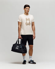 A man stands against a plain background wearing the Psycho Bunny MENS NICHOLAS EMBROIDERED TEE (MT0200054), black shorts, striped white socks, black Adidas sneakers, and holding a black duffel bag with a matching bunny logo.