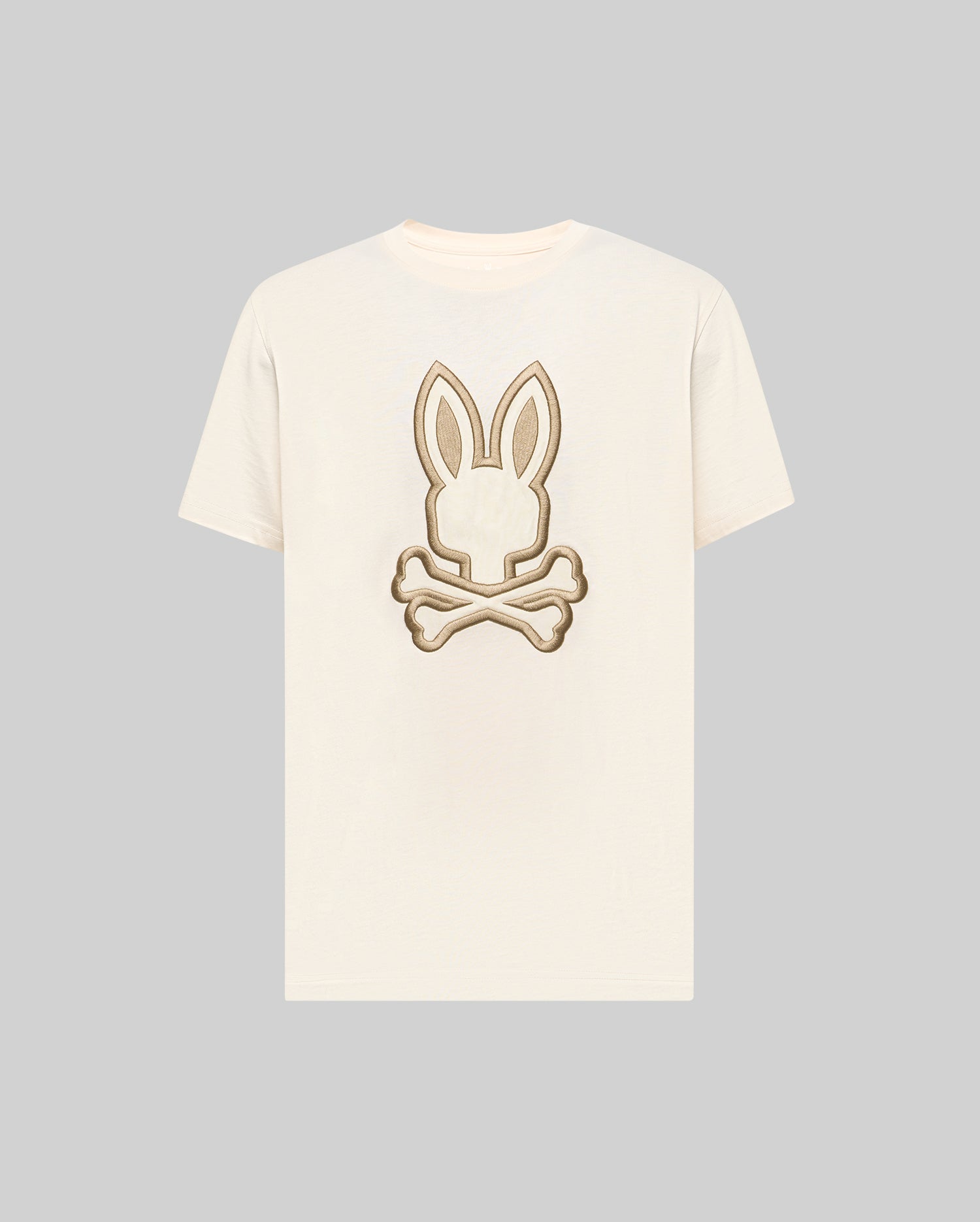 The Psycho Bunny MENS NICHOLAS EMBROIDERED TEE (MT0200054) features a cream base with a beige and brown embroidered bunny head and crossed bones motif centered on the chest.