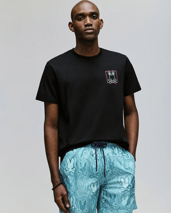 A man with a neutral expression stands against a plain background, wearing the Psycho Bunny MENS JESSE GRAPHIC TEE - MT0200056 in black and light blue bunny-patterned shorts, with his hands in his pockets.