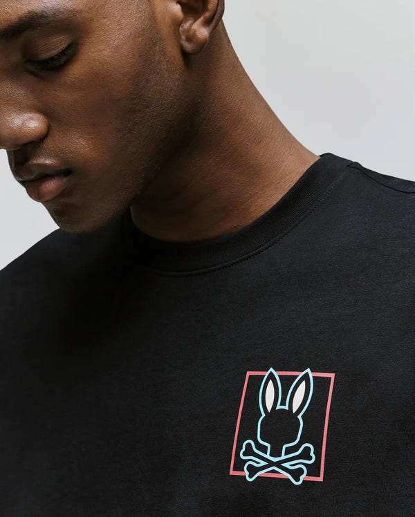 Wearing the Psycho Bunny MENS JESSE GRAPHIC TEE (MT0200056), a person looks down, partly obscuring their face. The black Pima cotton shirt features an eco-friendly water-based white bunny and crossbones graphic in a red square on the chest.