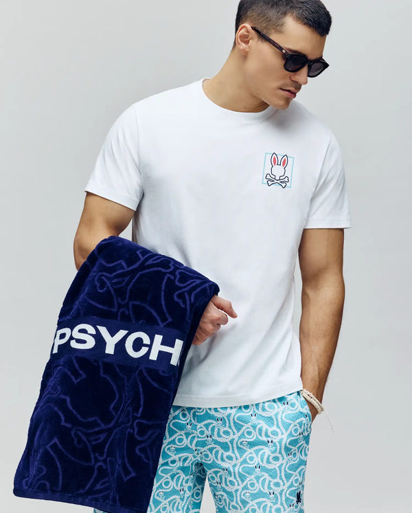 A man in sunglasses wears the Psycho Bunny MENS JESSE GRAPHIC TEE (MT0200056) with a bunny skull print and blue patterned shorts, holding a navy "PSYCH" towel. Water-based prints add an eco-friendly touch.