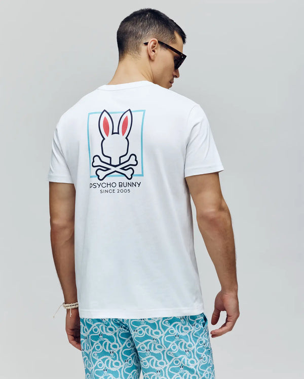 A man in Psycho Bunny's MENS JESSE GRAPHIC TEE (MT0200056), a white Pima cotton shirt with an eco-friendly logo on the back, blue patterned shorts, and sunglasses stands facing away against a plain background.