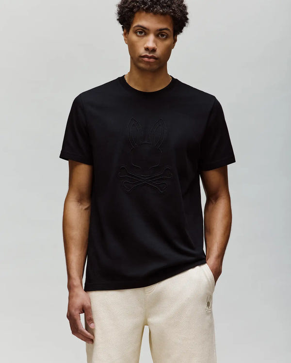 A man with short curly hair wears the Psycho Bunny MENS REMY GRAPHIC TEE (MT0200059) in black, made from soft Pima cotton with an embroidered Bunny logo and crossbones, paired with light beige pants against a plain light gray background.