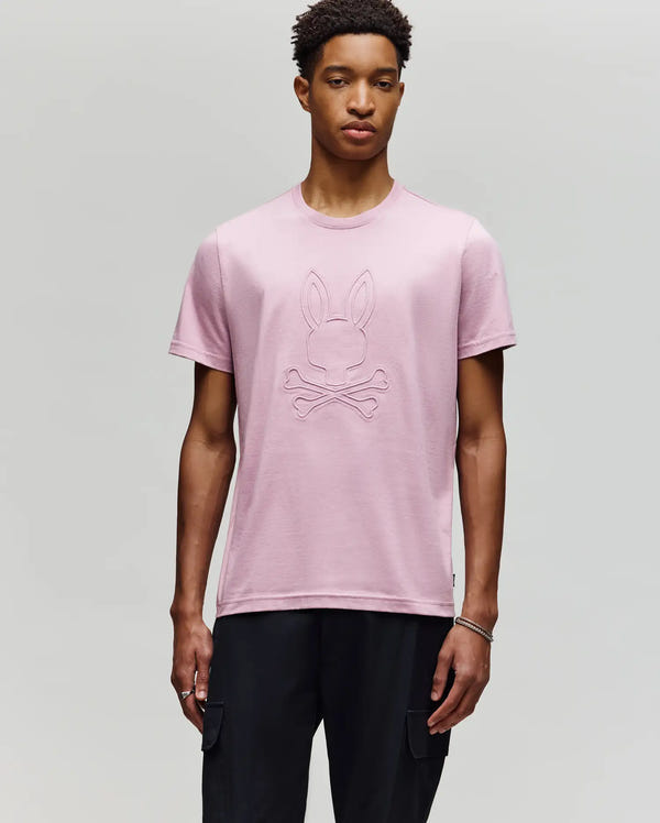 A young man with short curly hair and a neutral expression wears the Psycho Bunny MENS REMY GRAPHIC TEE - MT0200059 in light pink, made of soft Pima cotton with a raised bunny and crossbones graphic, paired with black pants against a plain background.