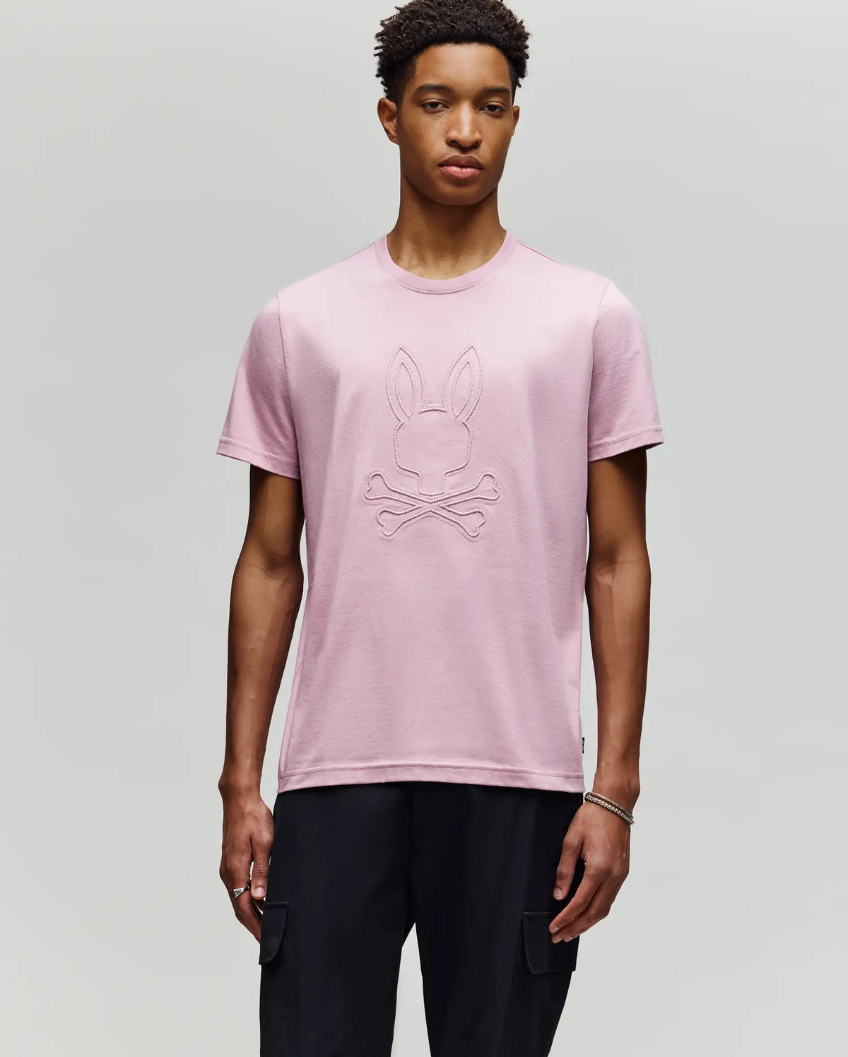 A young man with short curly hair and a neutral expression wears the Psycho Bunny MENS REMY GRAPHIC TEE - MT0200059 in light pink, made of soft Pima cotton with a raised bunny and crossbones graphic, paired with black pants against a plain background.