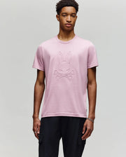 A young man with short curly hair and a neutral expression wears the Psycho Bunny MENS REMY GRAPHIC TEE - MT0200059 in light pink, made of soft Pima cotton with a raised bunny and crossbones graphic, paired with black pants against a plain background.