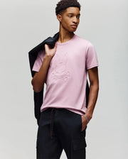 A man wears the Psycho Bunny MENS REMY GRAPHIC TEE - MT0200059 in light pink, featuring a bunny and crossbones logo in soft Pima cotton, paired with black pants. He has a black jacket over his shoulder and stands against a light gray background.