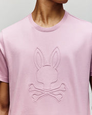 A person wears the Psycho Bunny MENS REMY GRAPHIC TEE (MT0200059) in pink Pima cotton, featuring the bunny logo embossed above two crossed bones on the chest. Their face is partially obscured.