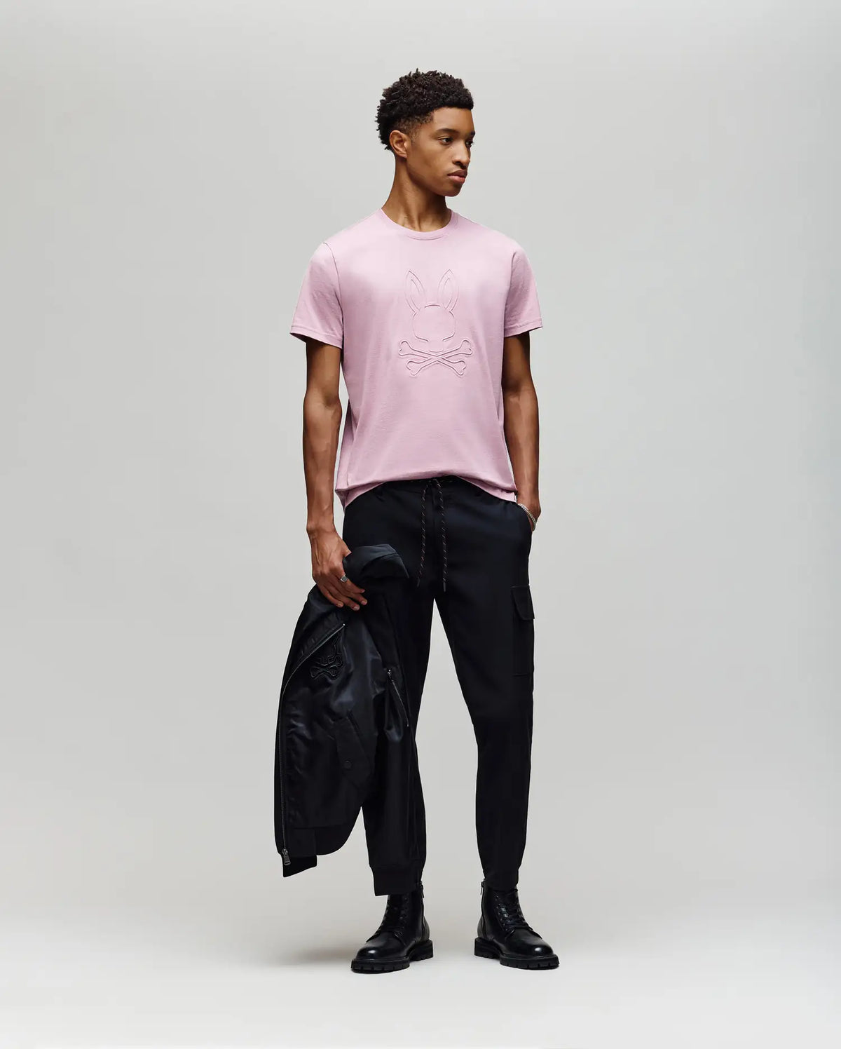 A man stands against a plain background wearing the Psycho Bunny MENS REMY GRAPHIC TEE - MT0200059 in light pink, paired with black cargo pants and boots. He holds a black jacket in one hand and looks to the side.