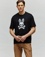 A man stands against a light gray background wearing the Psycho Bunny MENS RYDER OVERSIZED TEE (MT0200062), which features an embroidered Bunny skull and crossbones, paired with tan cargo pants.