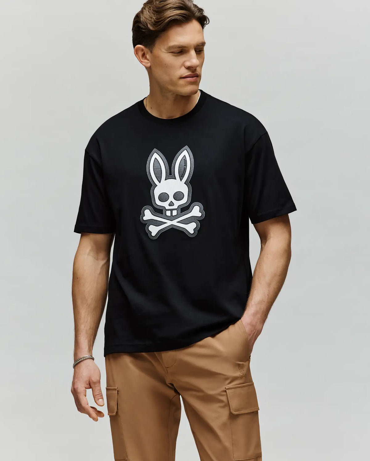 A man stands against a plain background, one hand in his tan cargo pants pocket, wearing the Psycho Bunny MENS RYDER OVERSIZED TEE (MT0200062), a premium black Pima cotton shirt with a white embroidered Bunny skull and crossbones.