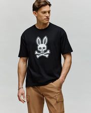 A man stands against a plain background, one hand in his tan cargo pants pocket, wearing the Psycho Bunny MENS RYDER OVERSIZED TEE (MT0200062), a premium black Pima cotton shirt with a white embroidered Bunny skull and crossbones.