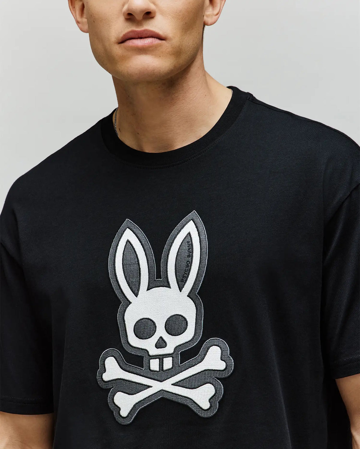 A person, face partially out of frame, wears the Psycho Bunny MENS RYDER OVERSIZED TEE (MT0200062), a premium Pima cotton shirt with a large skull and crossbones bunny ear graphic on the front.