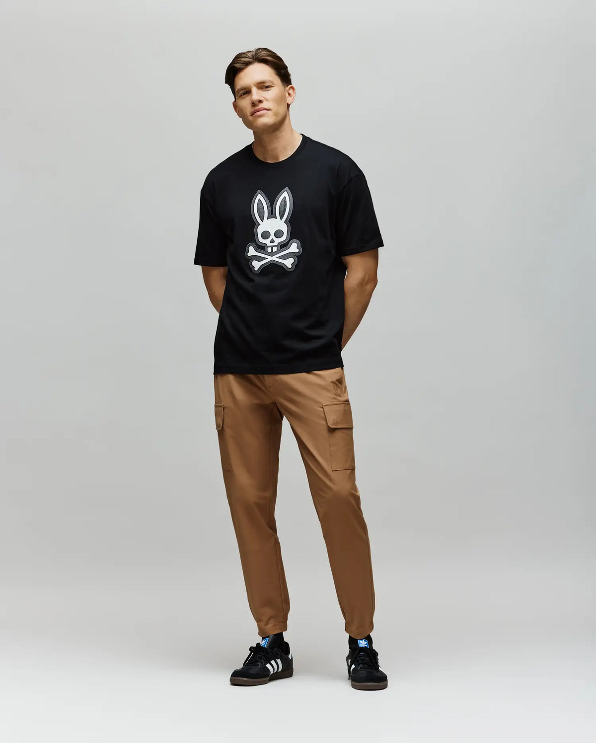 A man stands against a gray background, wearing the Psycho Bunny MENS RYDER OVERSIZED TEE (MT0200062) with an embroidered Bunny and crossbones graphic, tan cargo pants, and black Adidas sneakers. His hands are behind his back and he is smiling slightly.