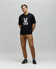 A man stands against a gray background, wearing the Psycho Bunny MENS RYDER OVERSIZED TEE (MT0200062) with an embroidered Bunny and crossbones graphic, tan cargo pants, and black Adidas sneakers. His hands are behind his back and he is smiling slightly.