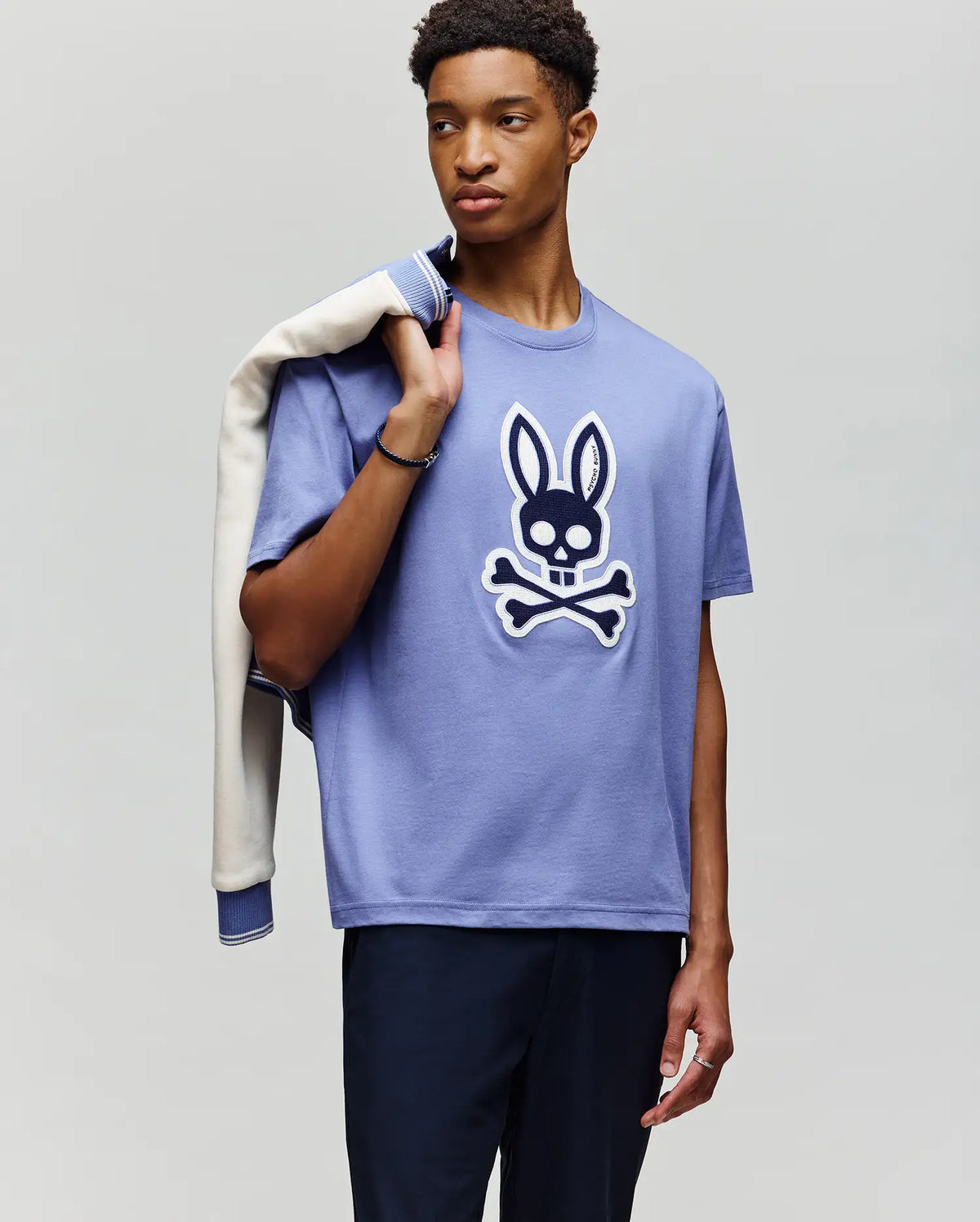 A young man stands holding a light jacket over his shoulder, wearing the Psycho Bunny MENS RYDER OVERSIZED TEE (MT0200062), a lavender Pima cotton t-shirt with a large embroidered Bunny skull-and-crossbones graphic and paired with dark pants.