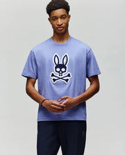 A young man with short curly hair wears the Psycho Bunny MENS RYDER OVERSIZED TEE (MT0200062), featuring a large bunny skull and crossbones graphic, standing against a plain light background, looking slightly downward with hands loosely clasped.