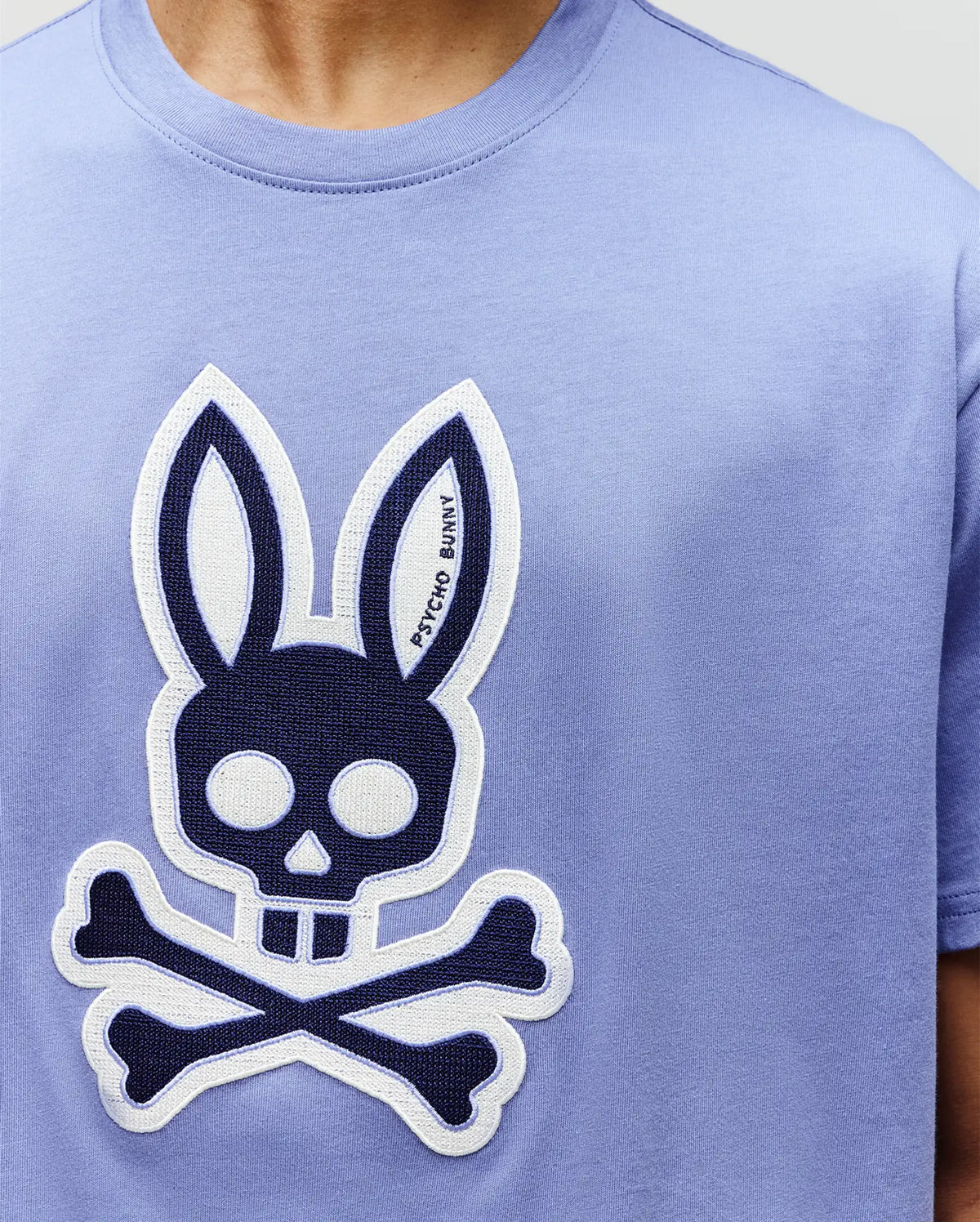 A person wearing the Psycho Bunny MENS RYDER OVERSIZED TEE (MT0200062), a light purple premium tee with a large embroidered Bunny skull and crossbones on the front, featuring the brand name in one ear.