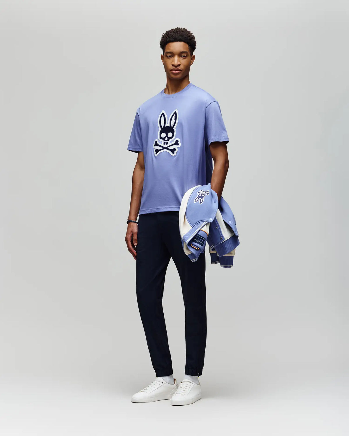 A man stands against a light gray background wearing the Psycho Bunny MENS RYDER OVERSIZED TEE (MT0200062) with an embroidered Bunny and crossbones, dark pants, white sneakers, and holding a matching jacket in his left hand.