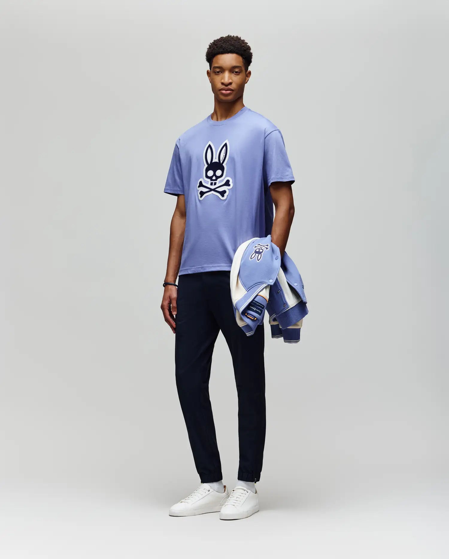 A man stands against a light gray background wearing the Psycho Bunny MENS RYDER OVERSIZED TEE (MT0200062) with an embroidered Bunny and crossbones, dark pants, white sneakers, and holding a matching jacket in his left hand.