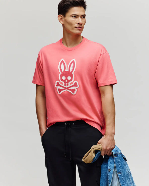 A man in the Psycho Bunny MENS RYDER OVERSIZED TEE (MT0200062), featuring premium Pima cotton and an embroidered Bunny and crossbones, stands with a denim jacket and beige cap against a plain background.