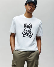 A person in the Psycho Bunny MENS RYDER OVERSIZED TEE (MT0200062)—a premium Pima cotton white T-shirt with a large black bunny skull and crossbones—olive cargo pants, and a black cap stands against a plain light gray background.