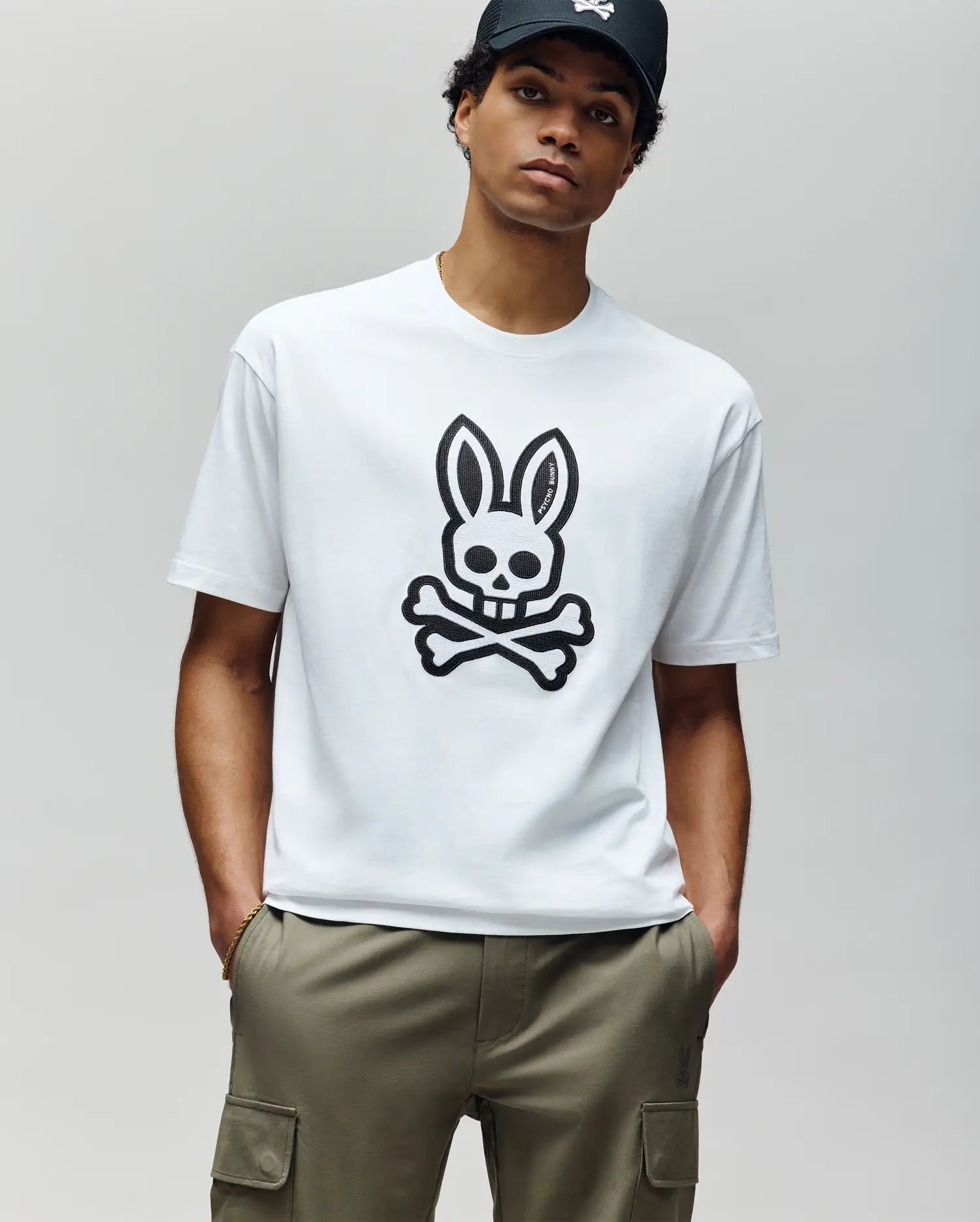 A person in the Psycho Bunny MENS RYDER OVERSIZED TEE (MT0200062)—a premium Pima cotton white T-shirt with a large black bunny skull and crossbones—olive cargo pants, and a black cap stands against a plain light gray background.