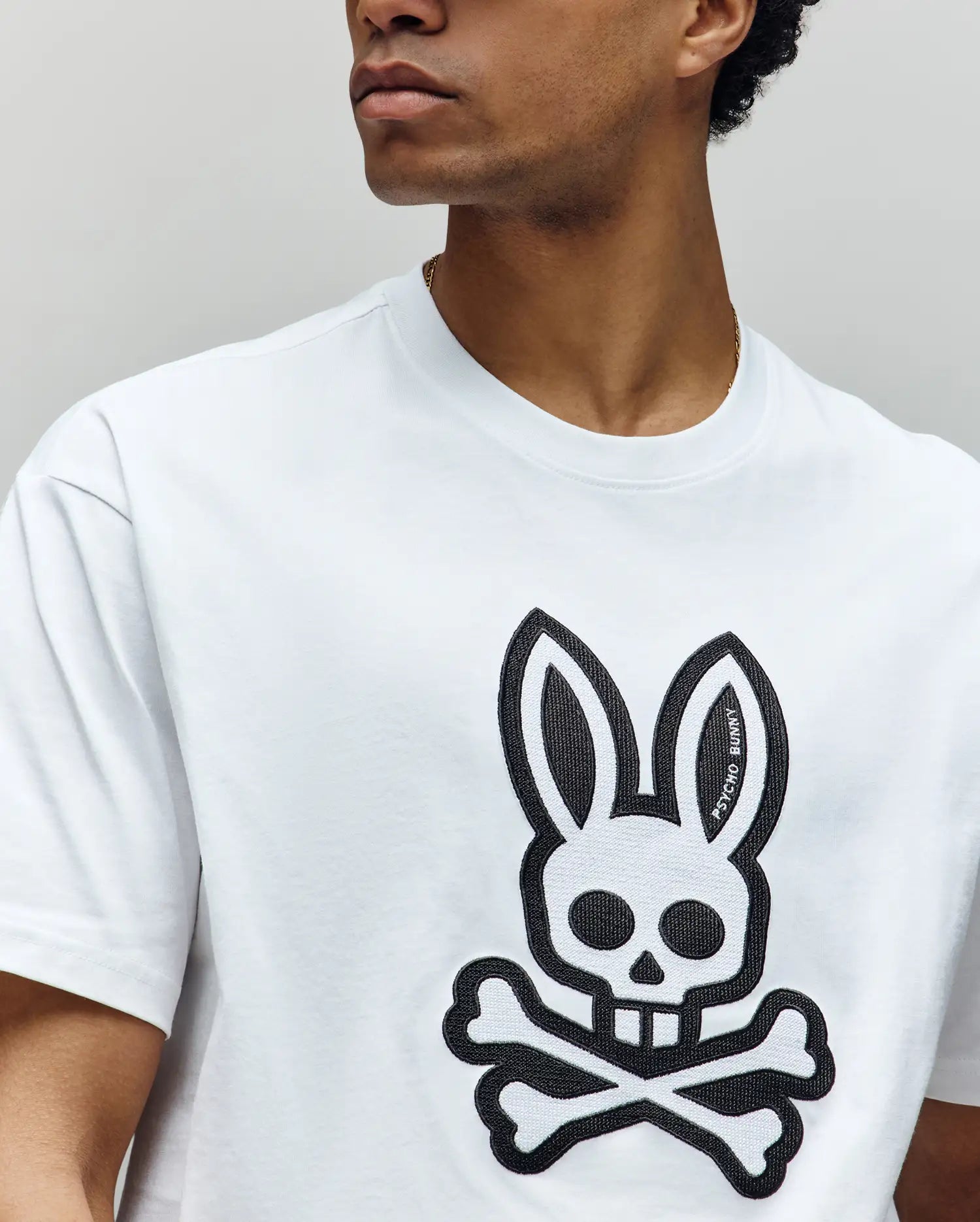 A person wears the Psycho Bunny MENS RYDER OVERSIZED TEE (MT0200062), a premium Pima cotton shirt with a large black skull and bunny ears graphic plus crossed bones on the front. The person's face is partially out of frame.