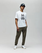A young man poses against a plain background wearing a black cap, Psycho Bunny MENS RYDER OVERSIZED TEE featuring a bold embroidered Bunny skull graphic, olive green joggers, and white sneakers.