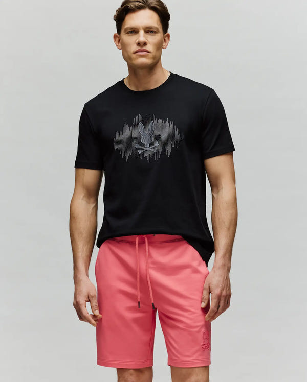 A man stands against a plain background wearing the Psycho Bunny MENS OLLIE GRAPHIC TEE (MT0200066) in black, featuring a white bunny graphic, and pink drawstring shorts, looking straight at the camera with a neutral expression.