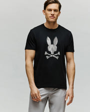 A man faces forward, wearing the Psycho Bunny MENS GRANT BUNNY GRAPHIC TEE - MT0200081 in black Pima cotton with a white striped bunny and crossbones HD graphic, paired with light gray pants against a light plain background.