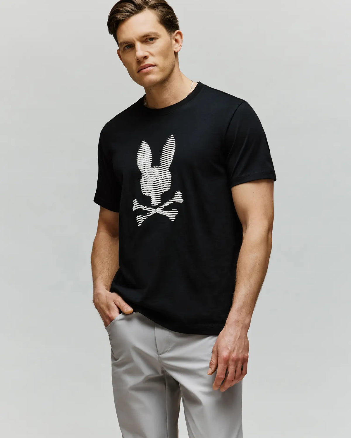 A man, wearing light gray pants and a Psycho Bunny MENS GRANT BUNNY GRAPHIC TEE (MT0200081) in soft black Pima cotton with an HD-printed white bunny skull and crossbones, stands with one hand in his pocket against a plain background.