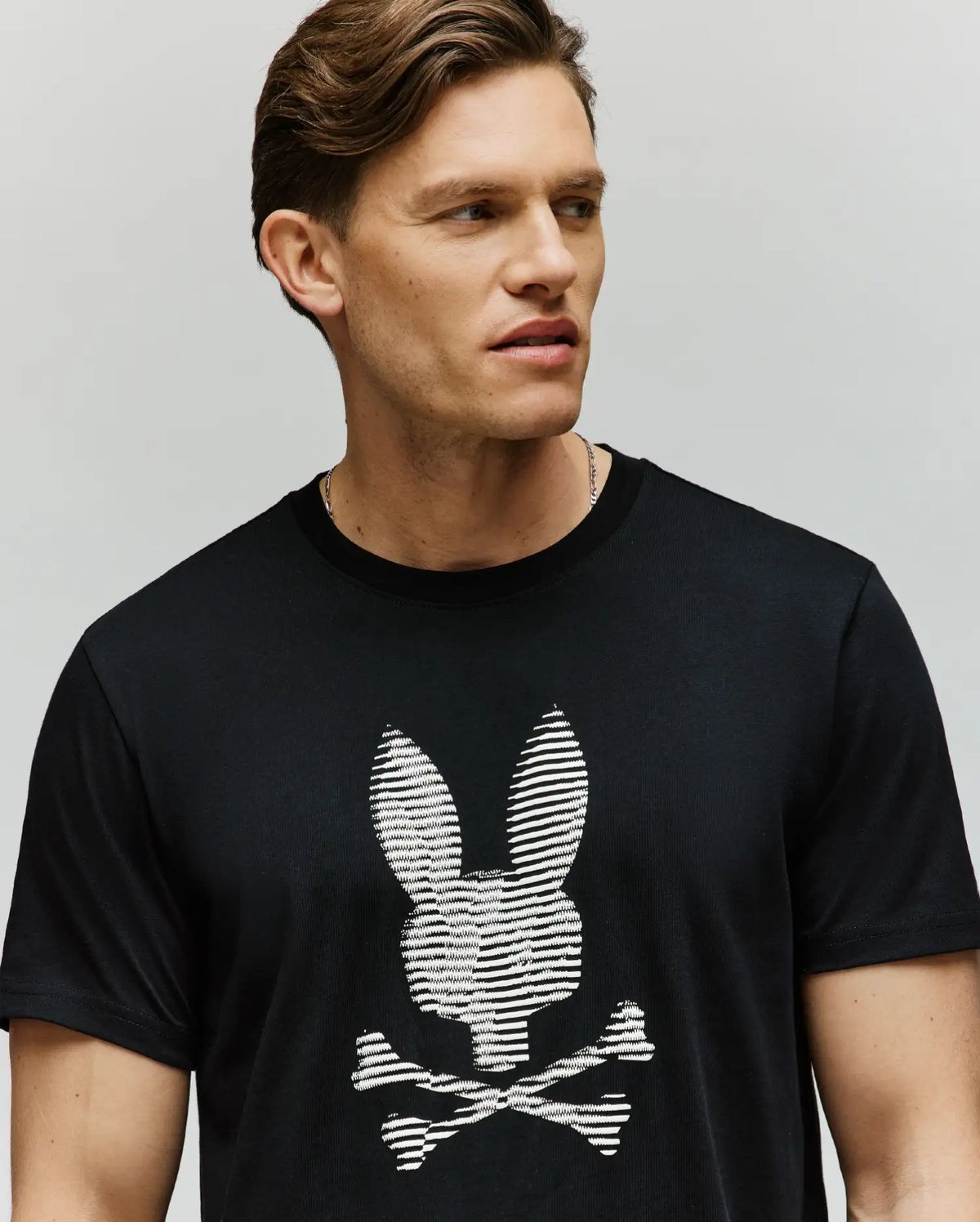 A man with short, styled brown hair models the Psycho Bunny MENS GRANT BUNNY GRAPHIC TEE (MT0200081), a black soft Pima cotton tee featuring an HD-printed white striped bunny head above crossed bones, set against a plain background.