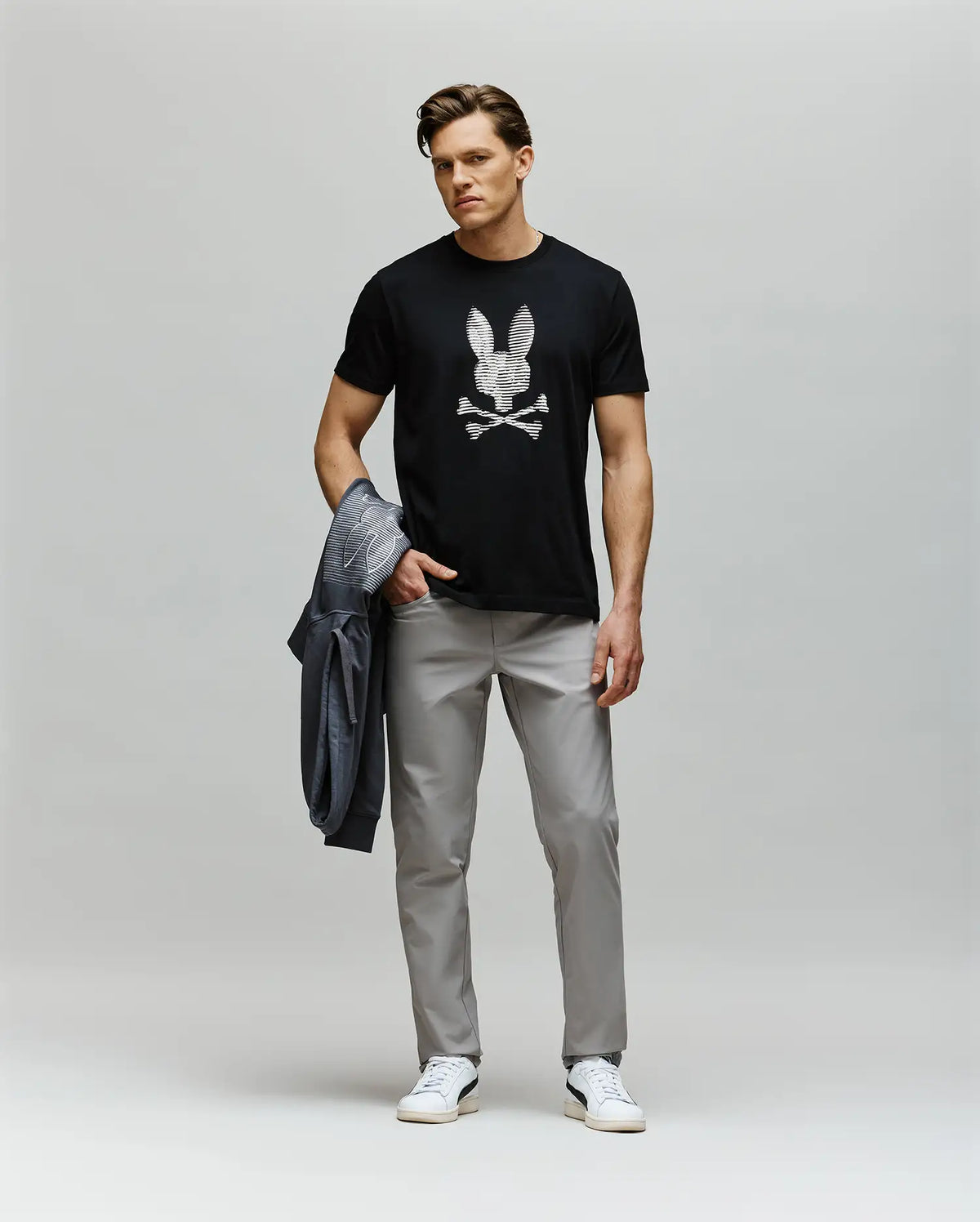 A man stands against a plain background wearing the Psycho Bunny MENS GRANT BUNNY GRAPHIC TEE (MT0200081) with an HD-printed skull-and-crossbones bunny logo, paired with gray pants and white sneakers. He holds a gray jacket in his left hand.