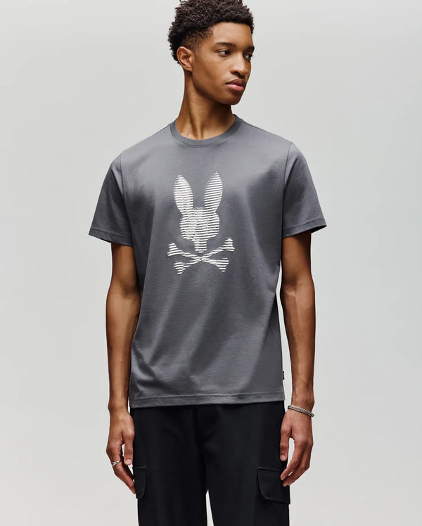 A young man wears the Psycho Bunny MENS GRANT BUNNY GRAPHIC TEE (MT0200081), a soft Pima cotton gray tee with a white bunny head and crossed bones, paired with black pants against a light background.