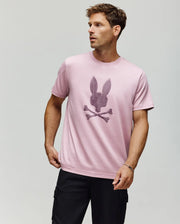 A man wears the Psycho Bunny MENS GRANT BUNNY GRAPHIC TEE (MT0200081) in light pink, made from soft Pima cotton in Peru, featuring a bold black bunny head and crossbones on the front, paired with black cargo pants against a plain background.