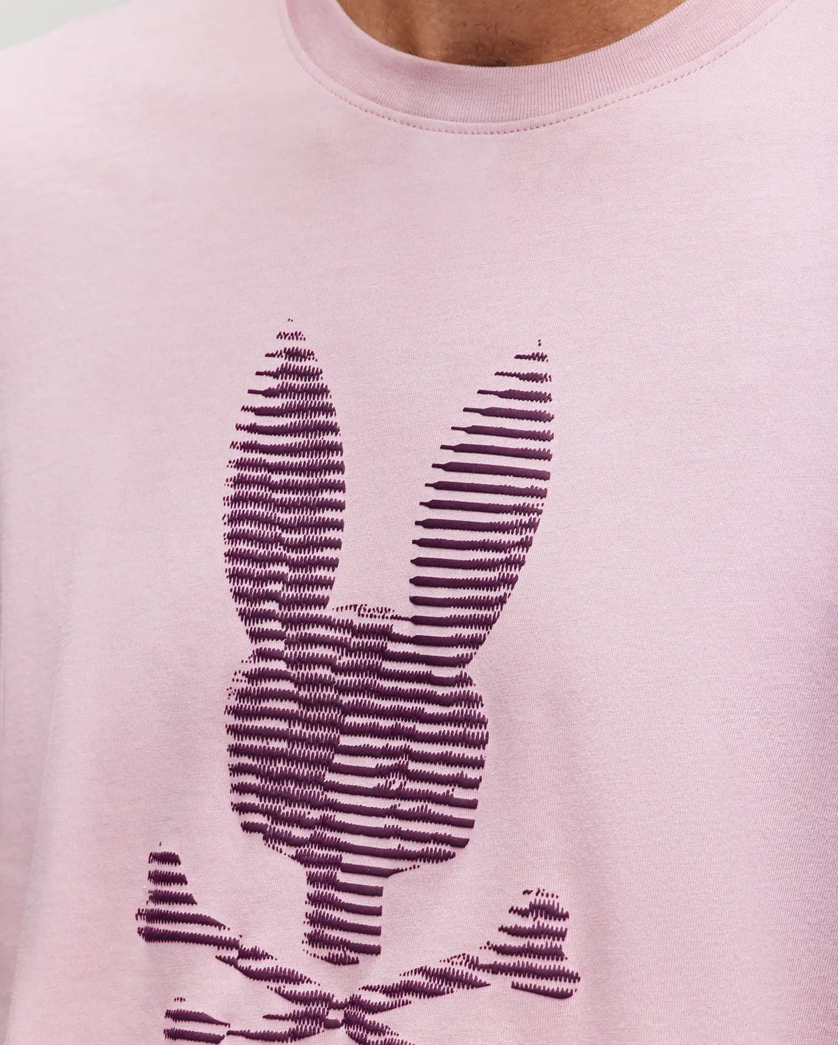 Close-up of the Psycho Bunny MENS GRANT BUNNY GRAPHIC TEE - MT0200081, made in Peru from soft Pima cotton in light pink, with a dark purple striped bunny head and crossed bones graphic on the chest.