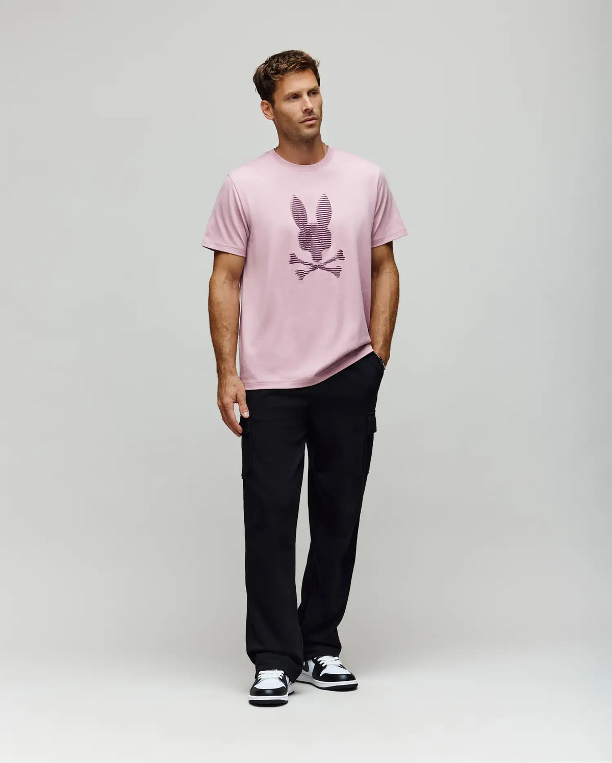 A man stands against a plain light background, wearing the Psycho Bunny MENS GRANT BUNNY GRAPHIC TEE (MT0200081), paired with black pants and black-and-white sneakers. His hands are in his pockets and he looks slightly to the side.