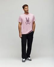 A man stands against a plain light background, wearing the Psycho Bunny MENS GRANT BUNNY GRAPHIC TEE (MT0200081), paired with black pants and black-and-white sneakers. His hands are in his pockets and he looks slightly to the side.