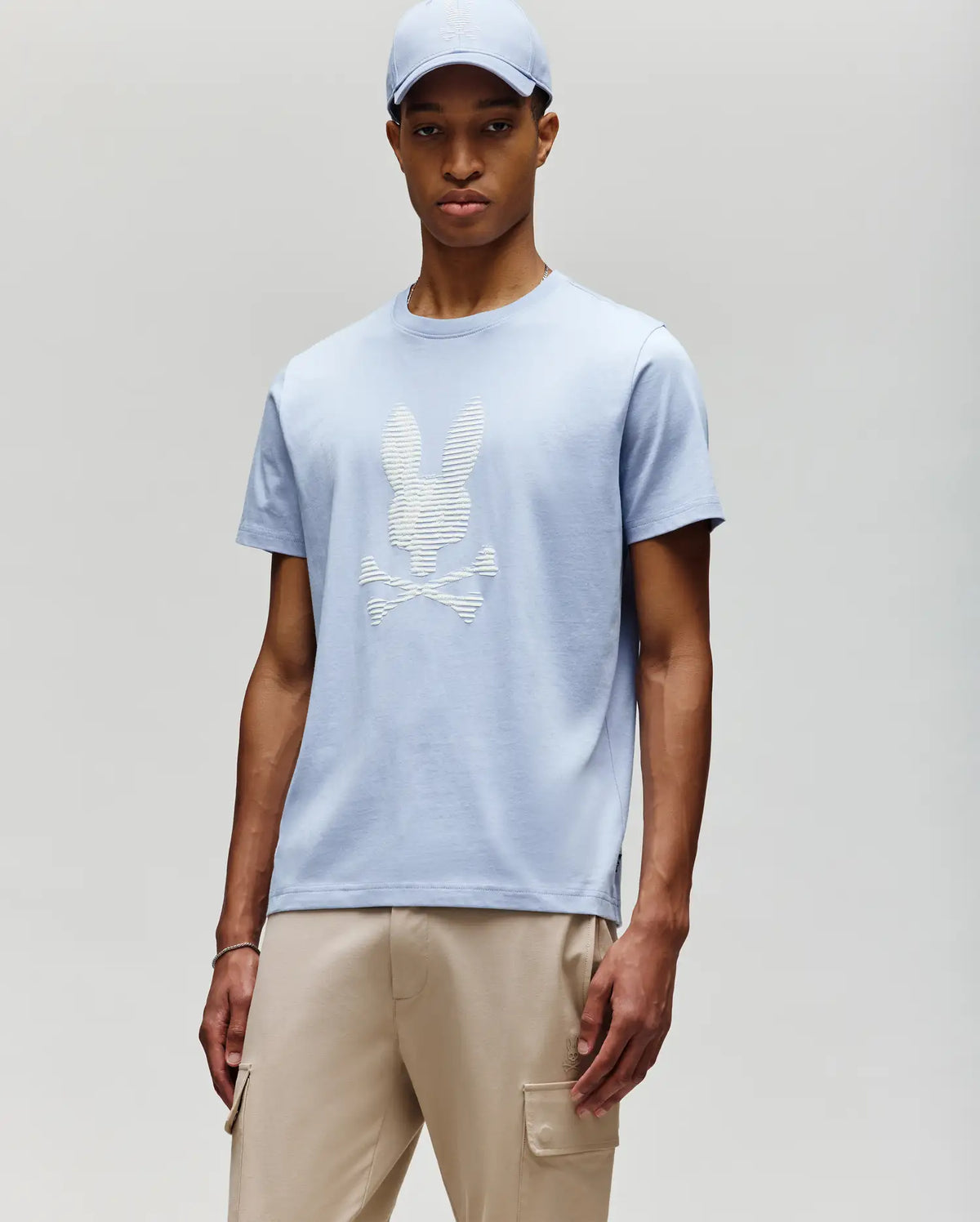 A man wears the Psycho Bunny MENS GRANT BUNNY GRAPHIC TEE - MT0200081 in light blue with a matching cap and beige cargo pants, standing against a neutral background for a fresh take on modern men's style.