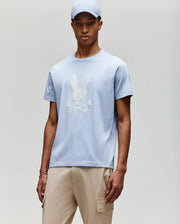 A man wears the Psycho Bunny MENS GRANT BUNNY GRAPHIC TEE - MT0200081 in light blue with a matching cap and beige cargo pants, standing against a neutral background for a fresh take on modern men's style.