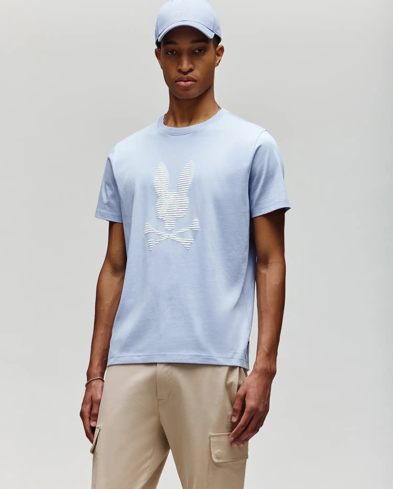 A man wears the Psycho Bunny MENS GRANT BUNNY GRAPHIC TEE - MT0200081 in light blue with a matching cap and beige cargo pants, standing against a neutral background for a fresh take on modern men's style.