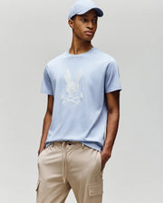 A person wears the Psycho Bunny MENS GRANT BUNNY GRAPHIC TEE - MT0200081 with a white skull and bunny logo, light blue cap, and beige drawstring pants against a plain backdrop—delivering fresh, modern men's style.