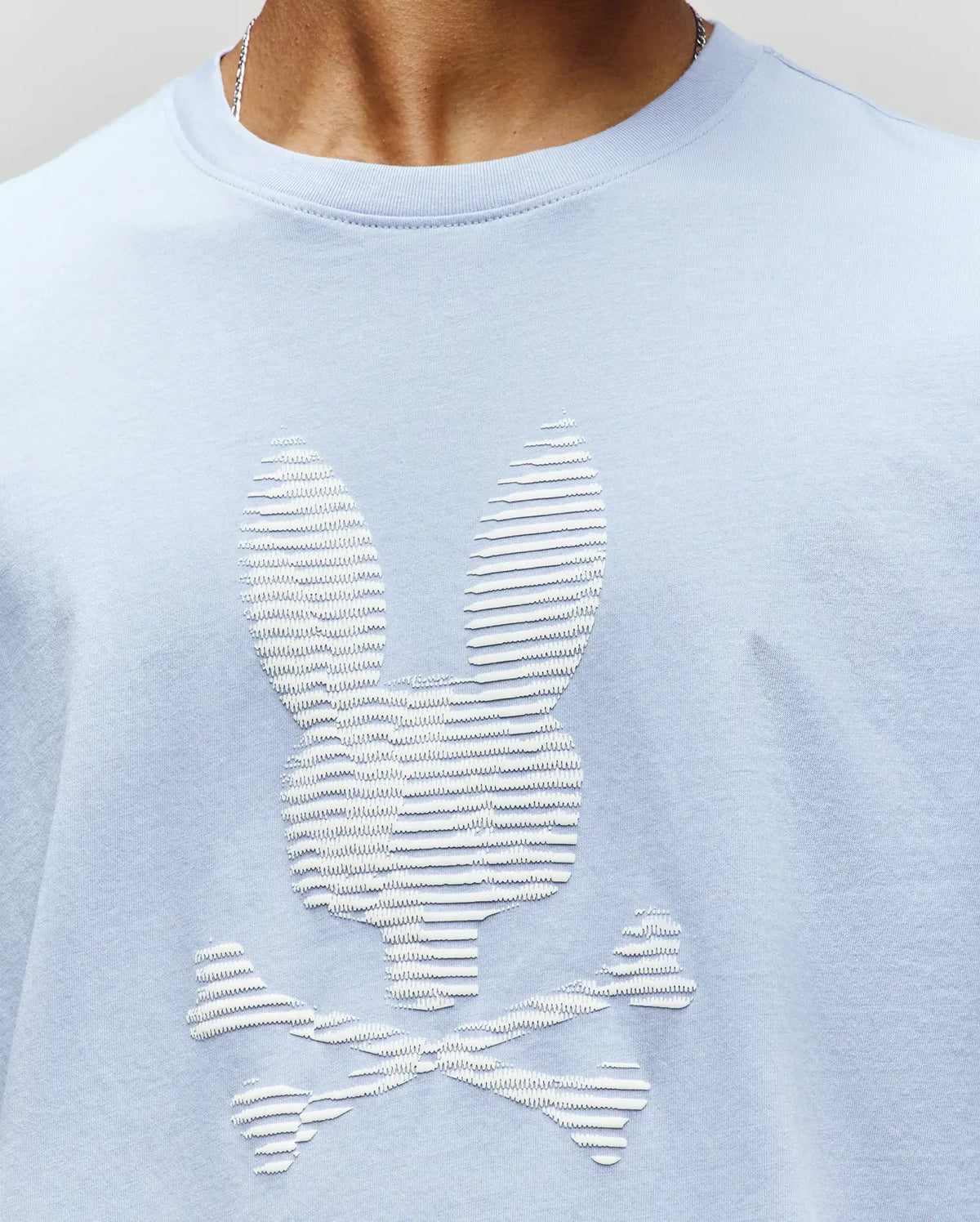 The Psycho Bunny MENS GRANT BUNNY GRAPHIC TEE (MT0200081) in light blue is crafted from soft Pima cotton and showcases a textured, raised bunny head above crossed bones for a playful touch on men’s fashion.