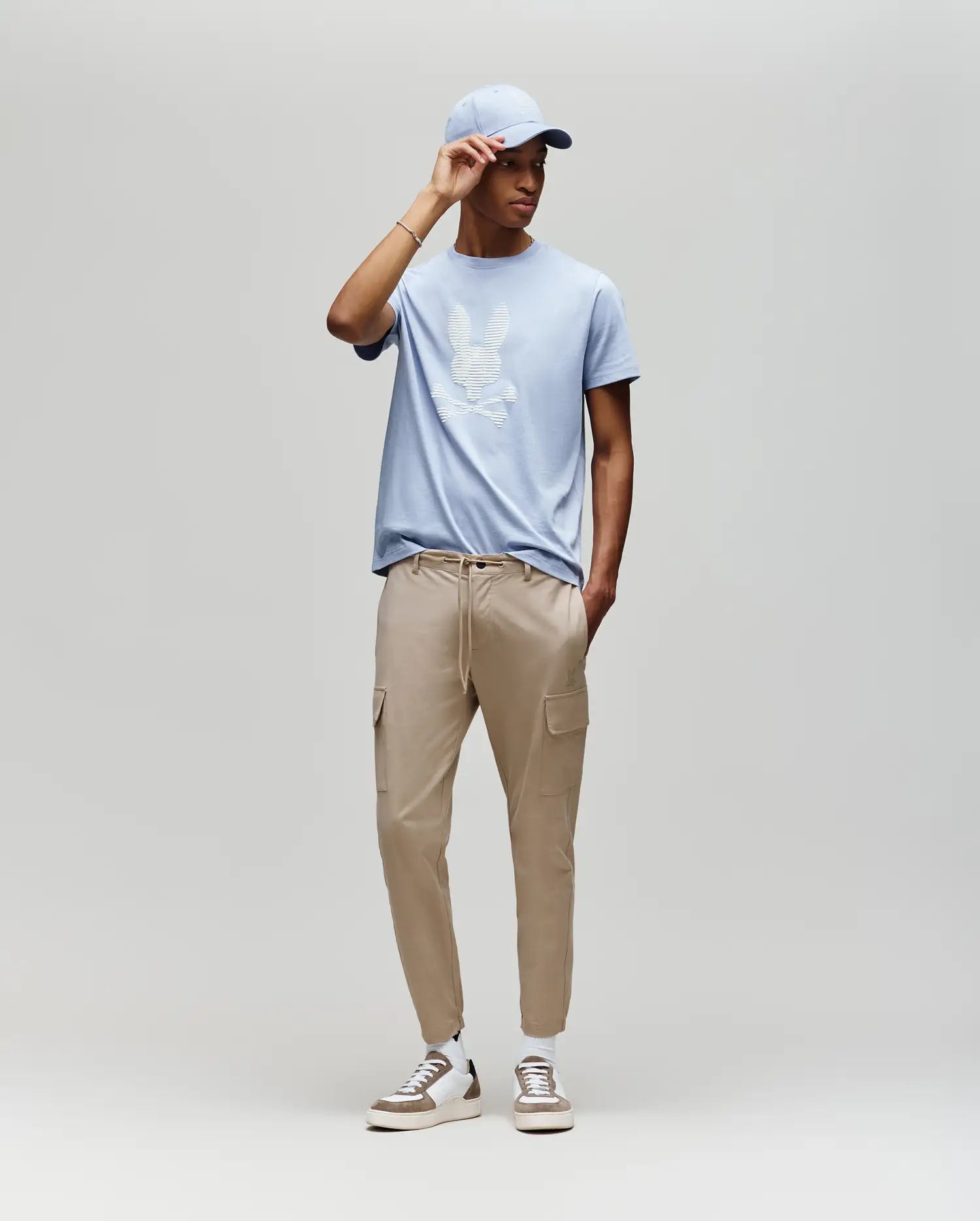 A man in a light blue Psycho Bunny MENS GRANT BUNNY GRAPHIC TEE (MT0200081), matching cap, beige cargo joggers, and beige sneakers stands against a plain gray background, highlighting modern men's style.