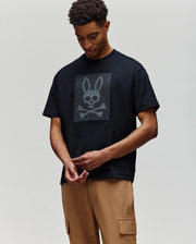 A person wears the Psycho Bunny MENS REECE OVERSIZED TEE (MT0200082), a black heavyweight jersey with a large bunny skull and crossbones graphic, paired with tan cargo pants, against a plain light background.