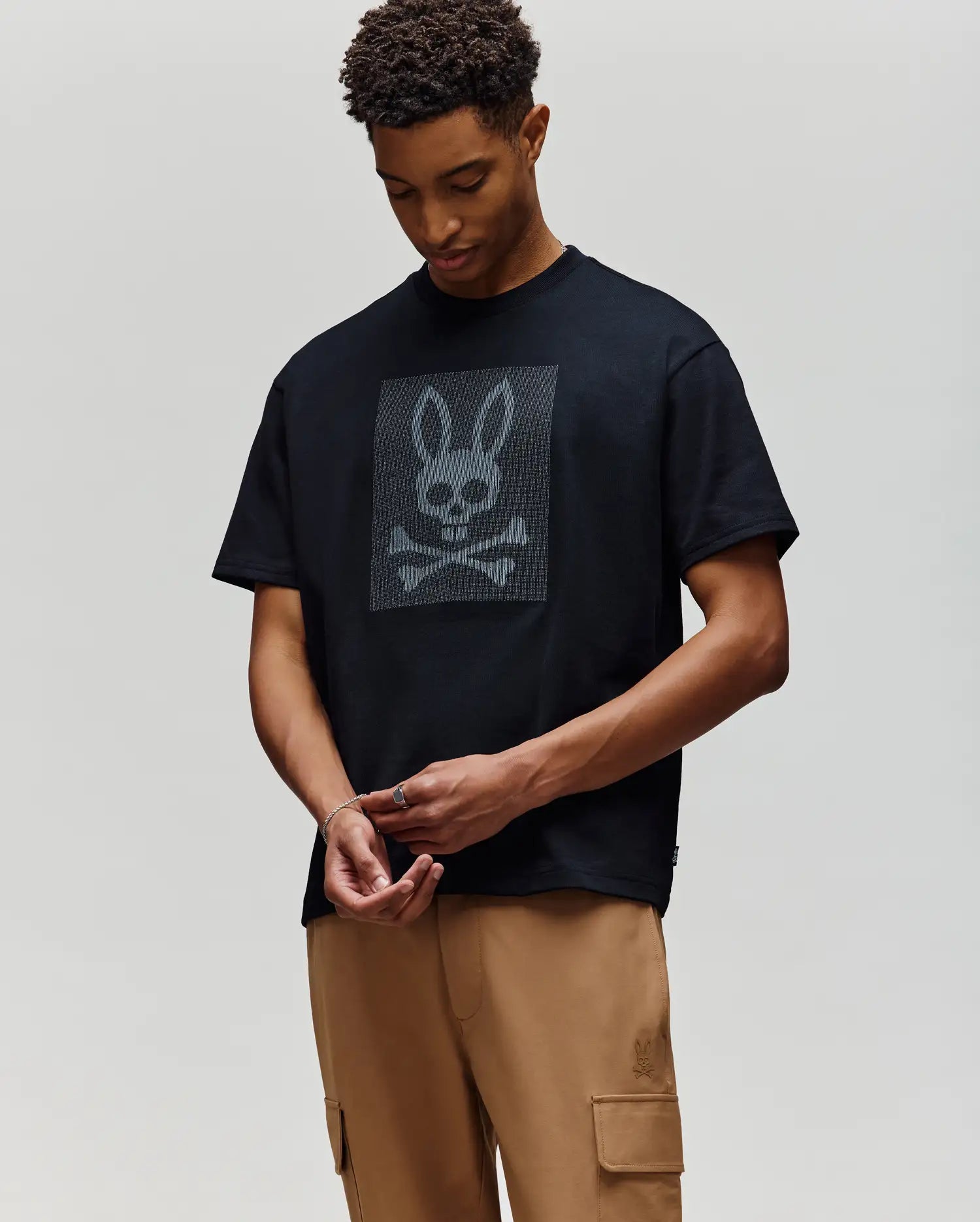 A person wears the Psycho Bunny MENS REECE OVERSIZED TEE (MT0200082), a black heavyweight jersey with a large bunny skull and crossbones graphic, paired with tan cargo pants, against a plain light background.