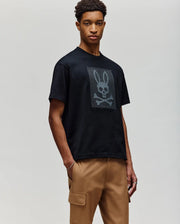 A man stands against a plain background wearing the Psycho Bunny MENS REECE OVERSIZED TEE (MT0200082) in heavyweight jersey, featuring a large bunny skull and crossbones on the chest, paired with tan cargo pants. His hands are in his pockets as he looks slightly aside.