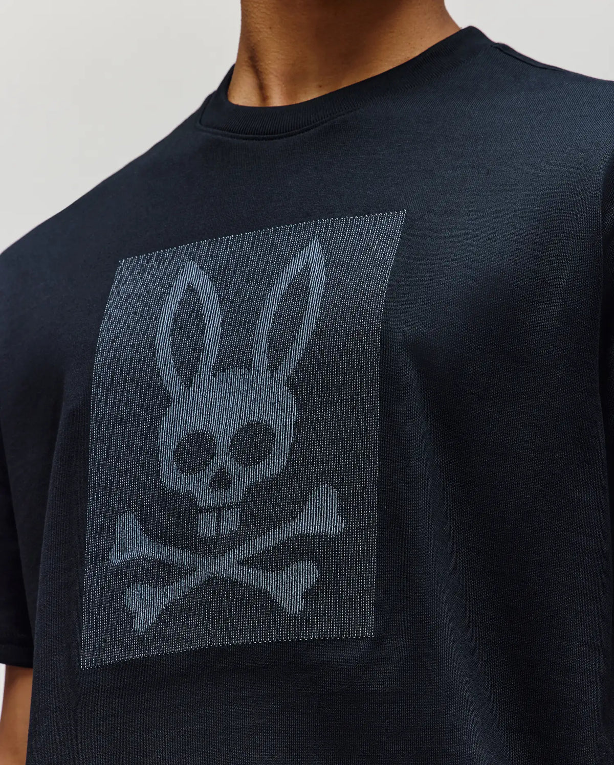 A person wears the Psycho Bunny MENS REECE OVERSIZED TEE (MT0200082), a black relaxed fit shirt featuring a chest graphic of a rabbit’s skull-like head with crossbones, printed in a rectangular area.