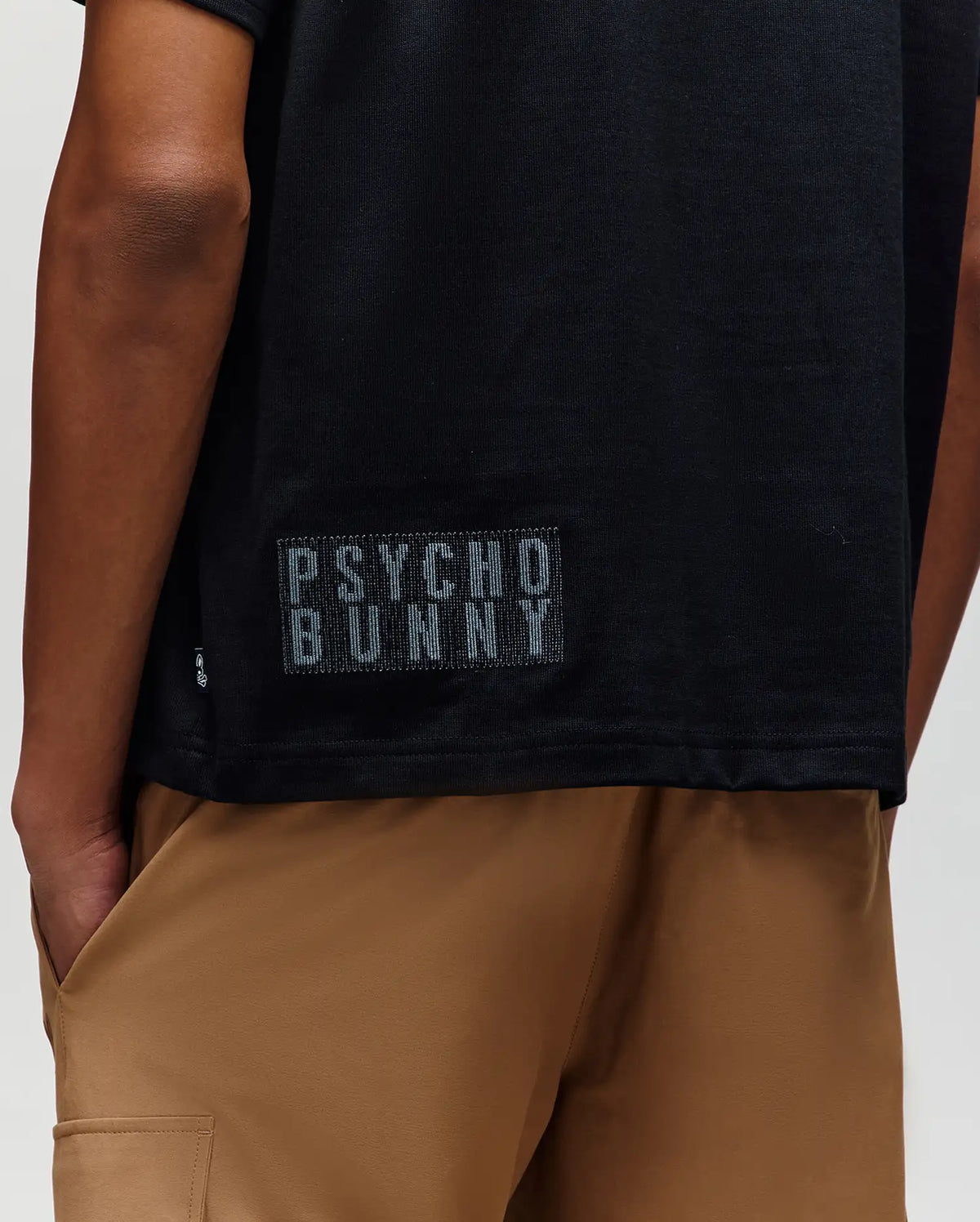 A person wearing the Psycho Bunny MENS REECE OVERSIZED TEE (MT0200082) in black, featuring a small "PSYCHO BUNNY" patch near the hem, paired with tan shorts; shown from mid-back to upper thigh.